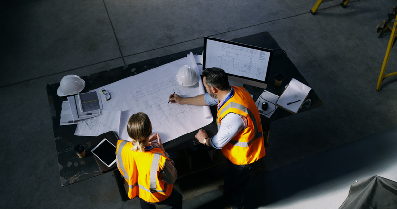 Vue du haut sur une table à dessin où deux ingénieurs vêtus d'un gilet orange travaillent sur un plan