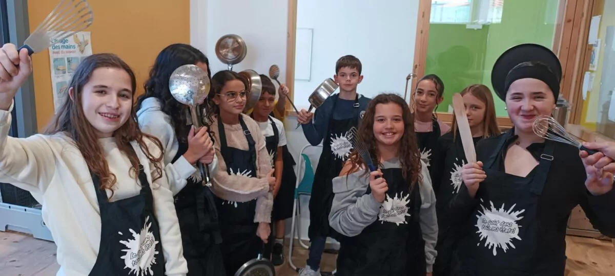 Des enfants vêtus de tabliers noirs brandissent des ustensiles de cuisine et s'apprêtent à cuisiner au sein de la cuisine du Pôle Jeunesse
