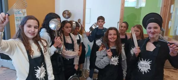 Des enfants vêtus de tabliers noirs brandissent des ustensiles de cuisine et s'apprêtent à cuisiner au sein de la cuisine du Pôle Jeunesse