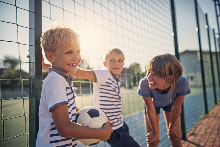 Trois enfants jouent au ballon en extérieur à proximité d'un stade