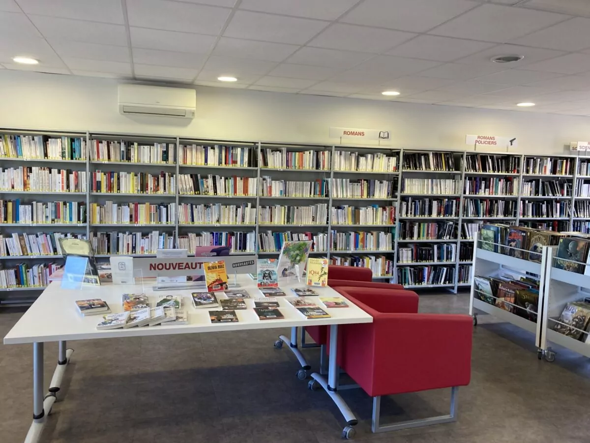 Vue intérieure de la bibliothèque avec des rayonnages de livres, une table présentant les nouveautés et des fauteuils rouges pour la lecture