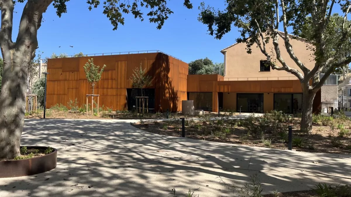 Bâtiment contemporain habillé de panneaux en acier Corten, intégré dans un espace paysager arboré avec une allée piétonne en béton et des plantations.