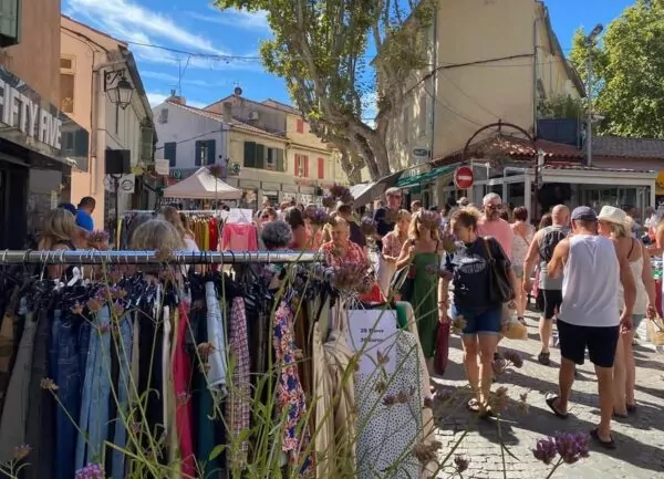 Marché en plein air dans un village provençal, avec des stands de vêtements, des commerces et de nombreux visiteurs sous les platanes.