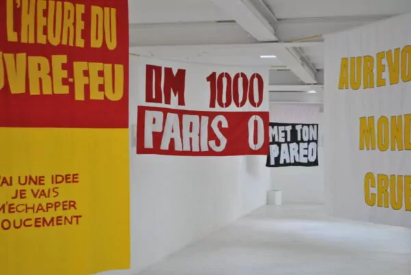 Vue d’une exposition consacrée aux textes et aux œuvres, présentant des affiches typographiques suspendues dans un espace blanc. Les compositions associent de grands lettrages rouges, jaunes et noirs à des expressions en français.