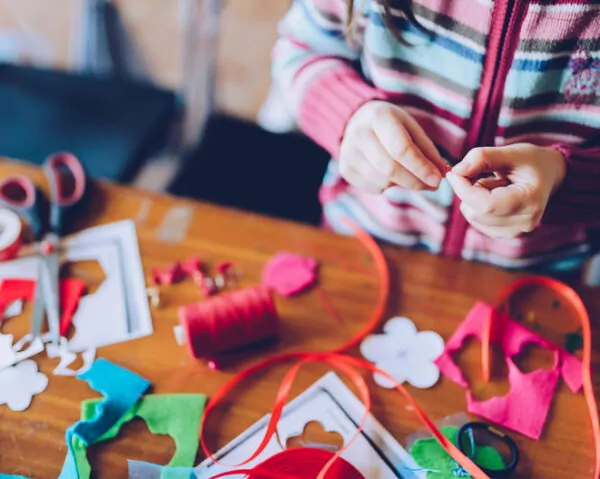 Mains d'enfant réalisant des créations manuelles avec des rubans, des fils colorés, du tissu et des accessoires disposés sur une table en bois.