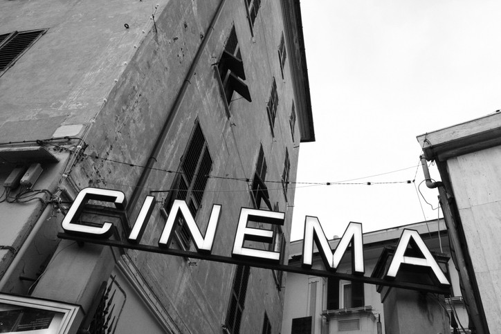Enseigne lumineuse « CINEMA » en noir et blanc, suspendue entre deux bâtiments anciens aux façades usées, avec des fenêtres ouvertes et des câbles électriques traversant la rue.
