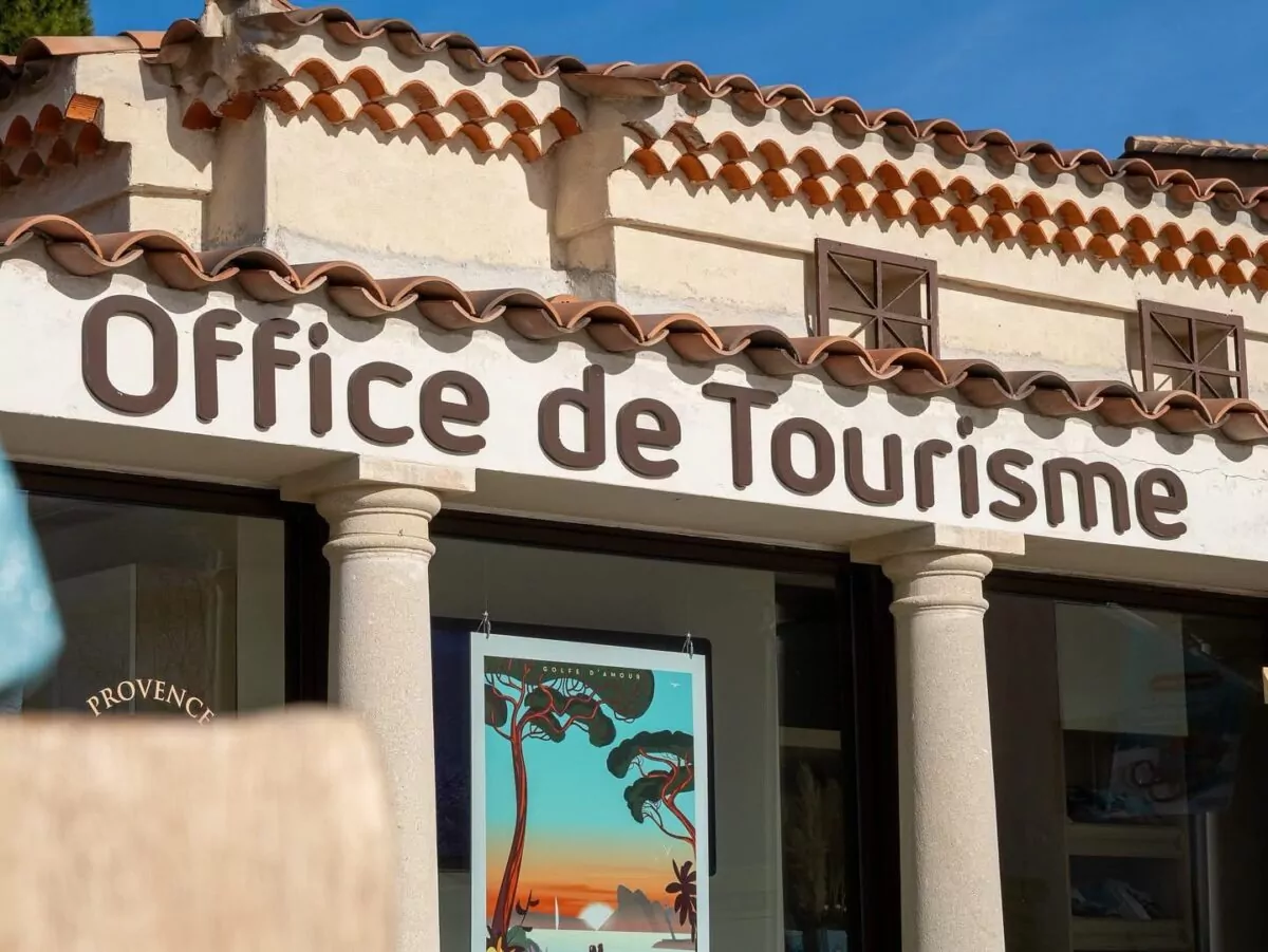 Façade de l’office de tourisme avec une enseigne en lettres brunes, des murs clairs, des tuiles provençales et une affiche colorée en vitrine, sous un ciel bleu ensoleillé.