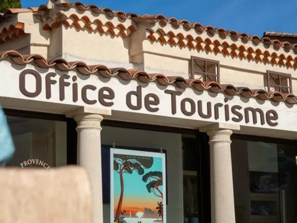 Façade de l’office de tourisme avec une enseigne en lettres brunes, des murs clairs, des tuiles provençales et une affiche colorée en vitrine, sous un ciel bleu ensoleillé.